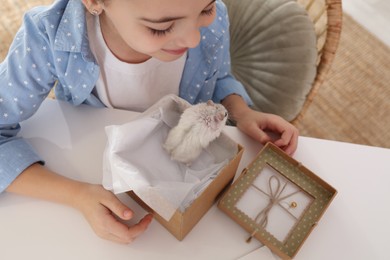 Happy little girl holding gift box with cute hamster at home, above view