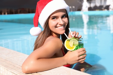 Photo of Young woman wearing Santa Claus hat with refreshing drink in swimming pool. Christmas vacation