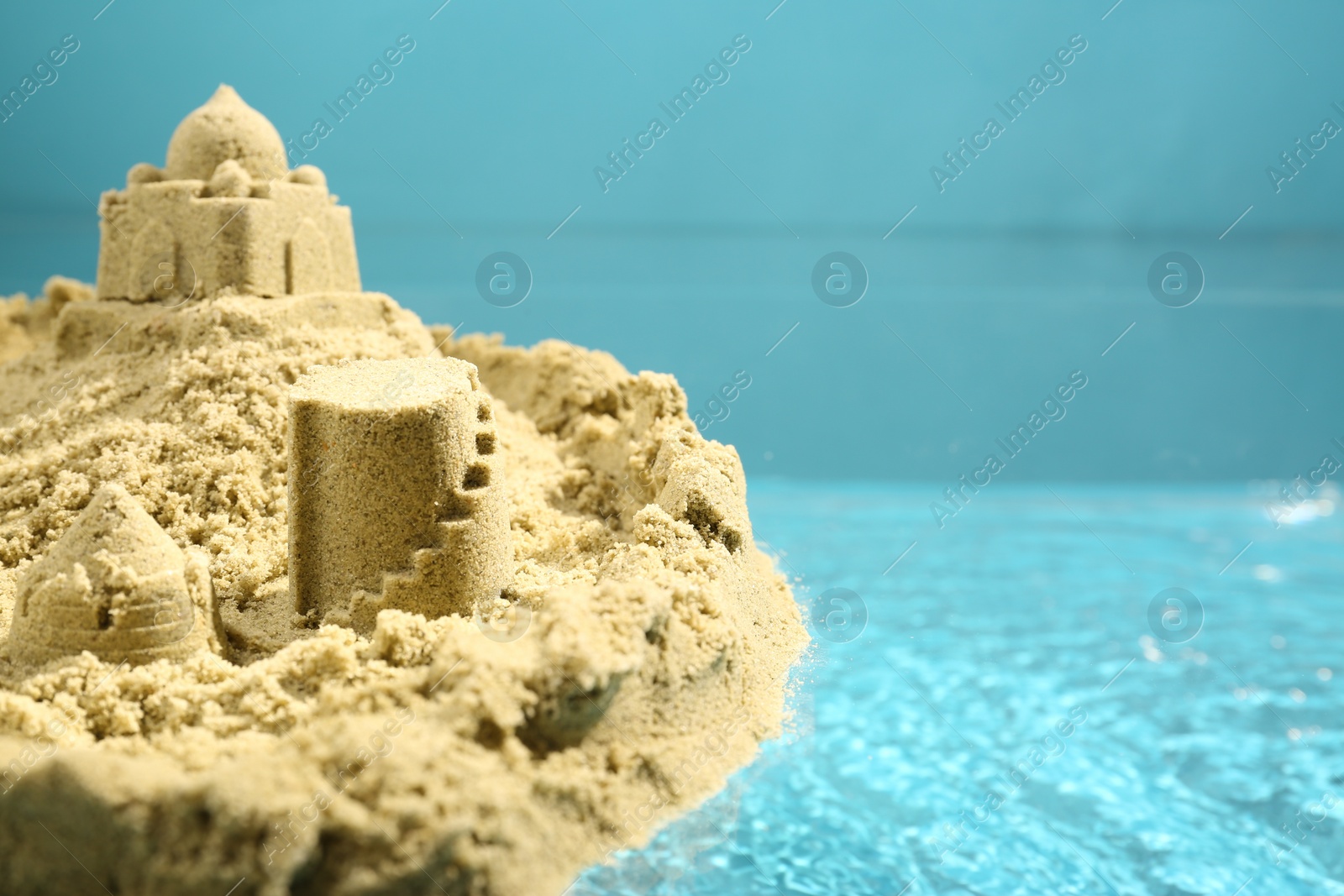 Photo of Pile of sand with beautiful castle on rippled water against light blue background, closeup. Space for text