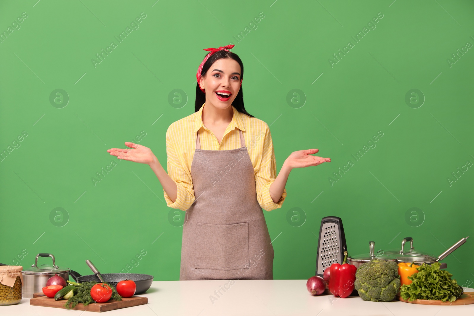 Photo of Emotional housewife at white table with vegetables and different utensils on green background