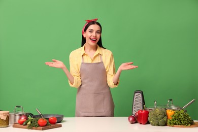 Emotional housewife at white table with vegetables and different utensils on green background