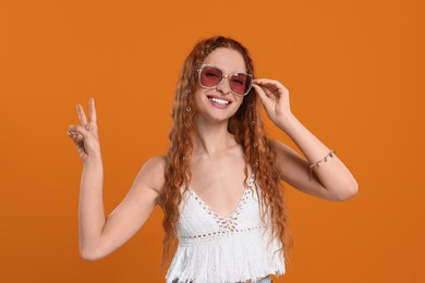 Photo of Stylish young hippie woman in sunglasses showing V-sign on orange background