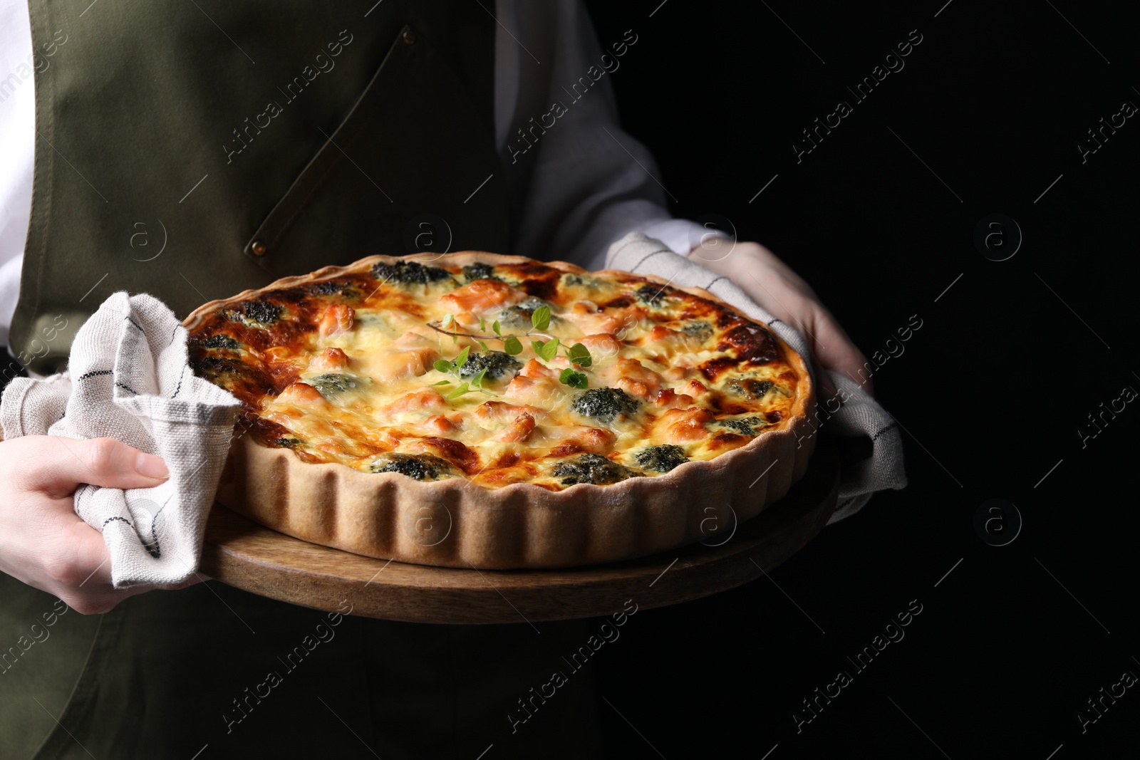 Photo of Woman holding wooden board with delicious homemade salmon quiche on black background, closeup