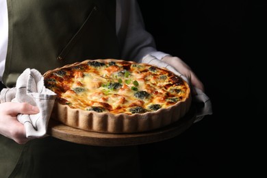 Woman holding wooden board with delicious homemade salmon quiche on black background, closeup