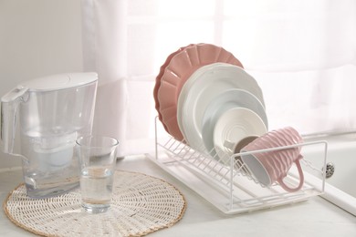 Photo of Drainer with different clean dishware, cup, glass and filter jug on light table indoors