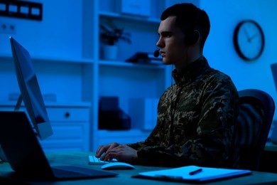 Image of Military service. Soldier in headphones working on computer at table in office at night