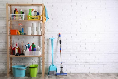 Photo of Shelving unit with detergents, cleaning tools and toilet paper near white brick wall indoors. Space for text