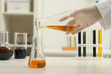 Photo of Laboratory worker pouring orange crude oil into flask at light marble table, closeup
