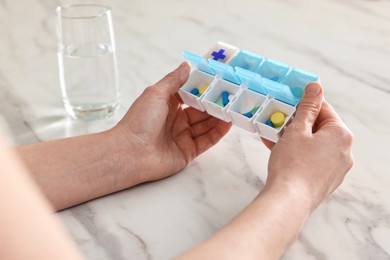 Woman with pills, organizer and glass of water at white marble table, closeup