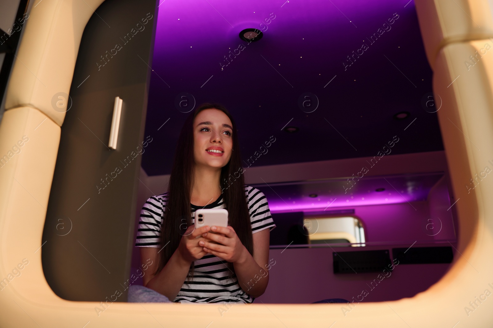 Photo of Happy young woman using smartphone in capsule of pod hostel