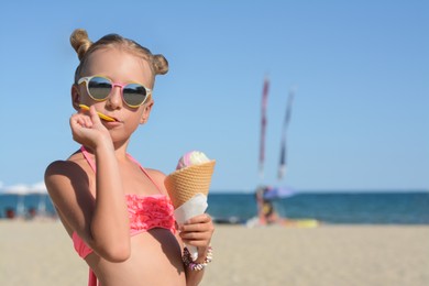 Photo of Adorable little girl in swimsuit with delicious ice cream at beach on sunny summer day, space for text