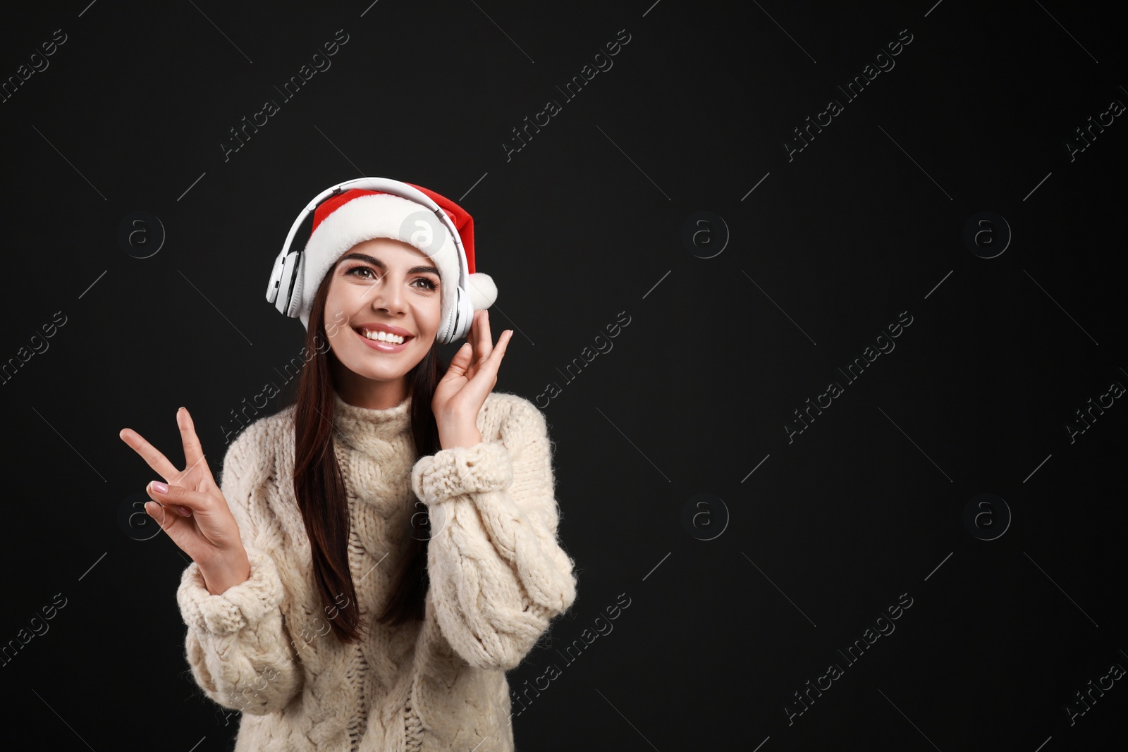 Photo of Young woman in Santa hat listening to Christmas music on black background, space for text