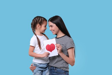 Happy woman with her cute daughter and handmade greeting card on light blue background. Mother's day celebration