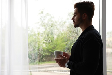 Photo of Thoughtful handsome man with cup of coffee near window indoors on rainy day