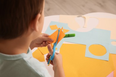 Photo of Little boy cutting color paper with scissors at table indoors, closeup