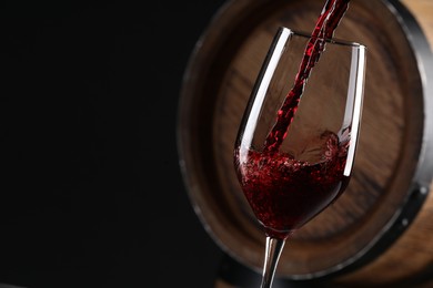 Photo of Pouring red wine into glass near wooden barrel against black background, closeup. Space for text
