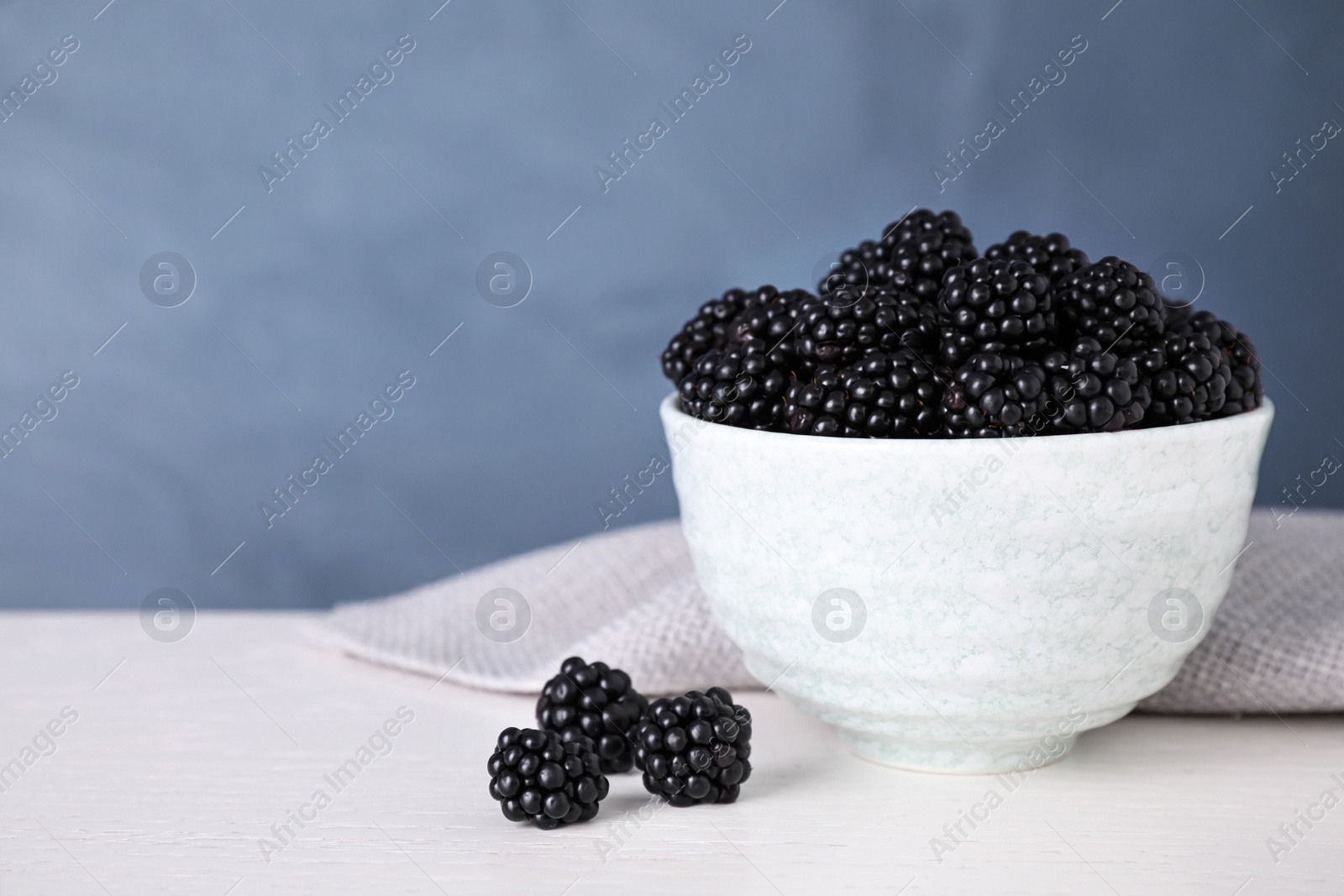 Photo of Fresh ripe blackberries in bowl on white table against blue background. Space for text