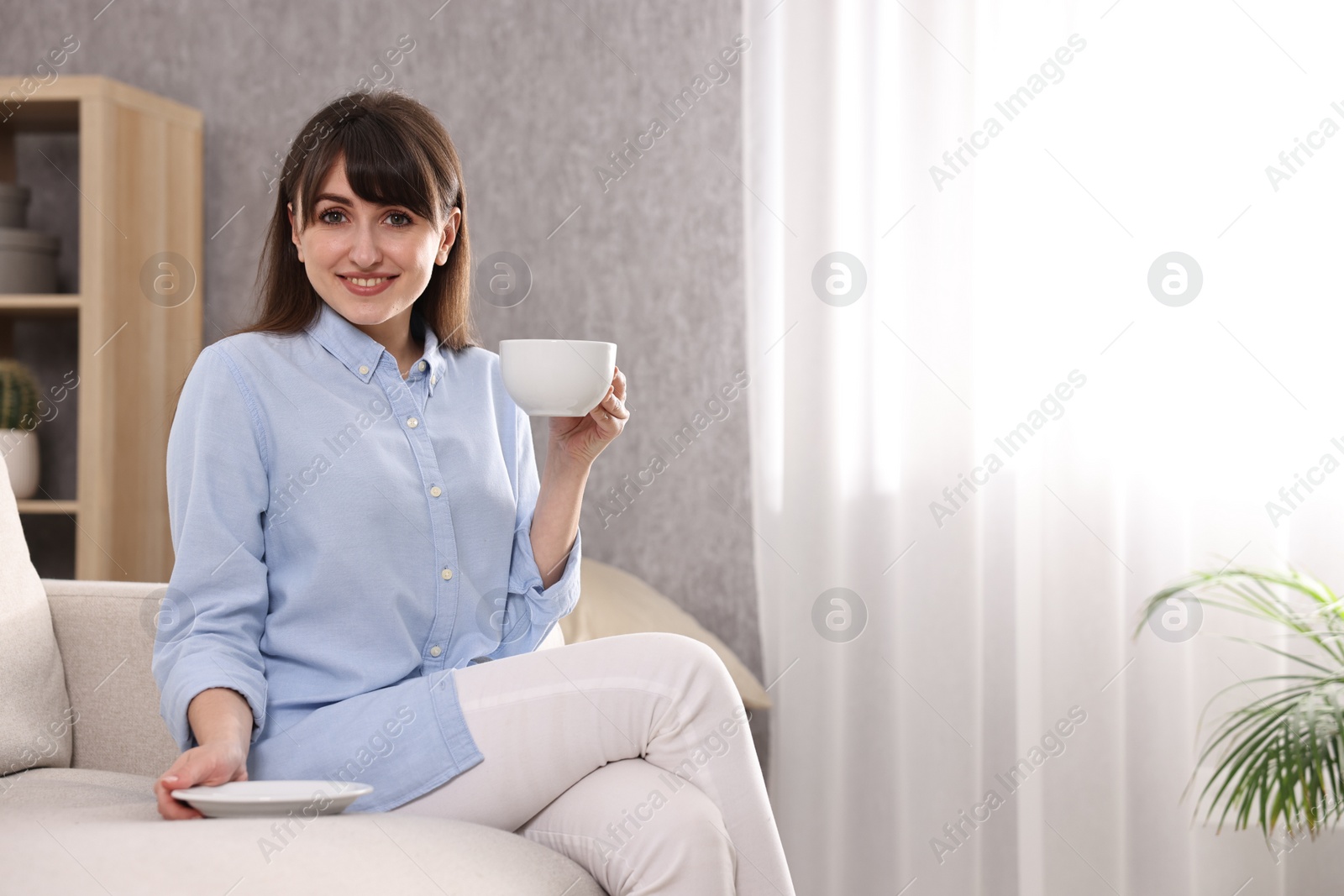 Photo of Beautiful young housewife with cup of drink on sofa at home, space for text