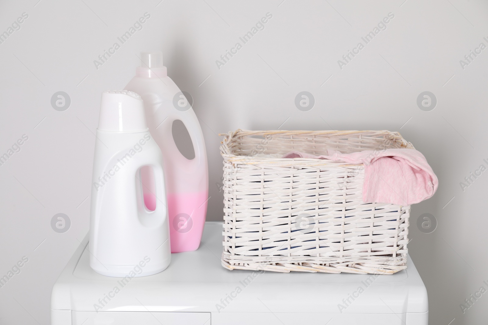 Photo of Baby clothes in wicker basket and laundry detergents on washing machine near light wall