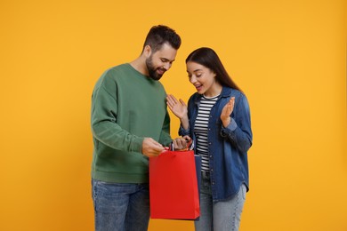 Photo of Man showing shopping bag with purchase to his excited girlfriend on orange background