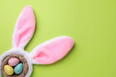 Photo of Headband with Easter bunny ears and dyed eggs in nest on green background, flat lay. Space for text