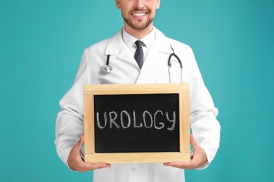 Male doctor holding small blackboard with word UROLOGY on turquoise background, closeup