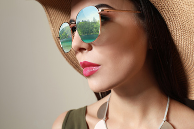 Young woman wearing stylish sunglasses with reflection of lake and hat on beige background 