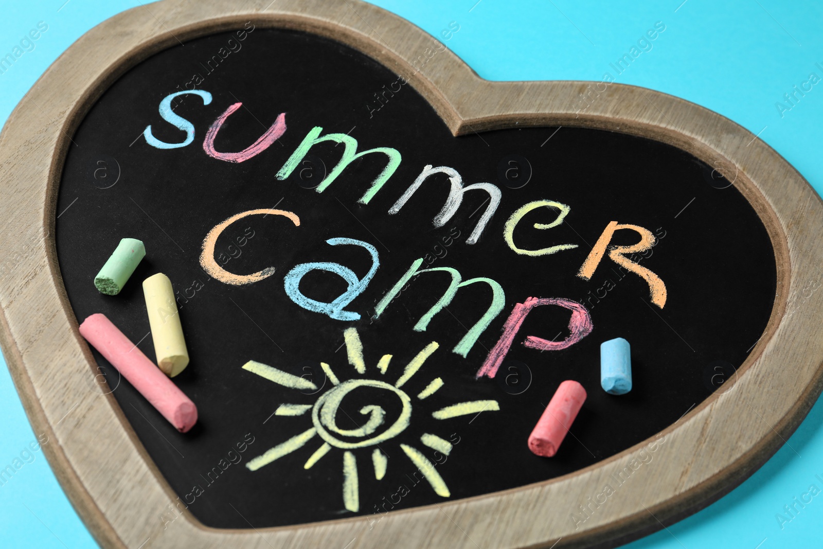 Photo of Heart shaped blackboard with text SUMMER CAMP, drawing and chalk sticks on color background