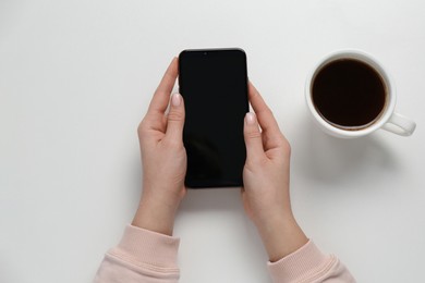 Photo of Woman with smartphone and cup of coffee at white table, top view