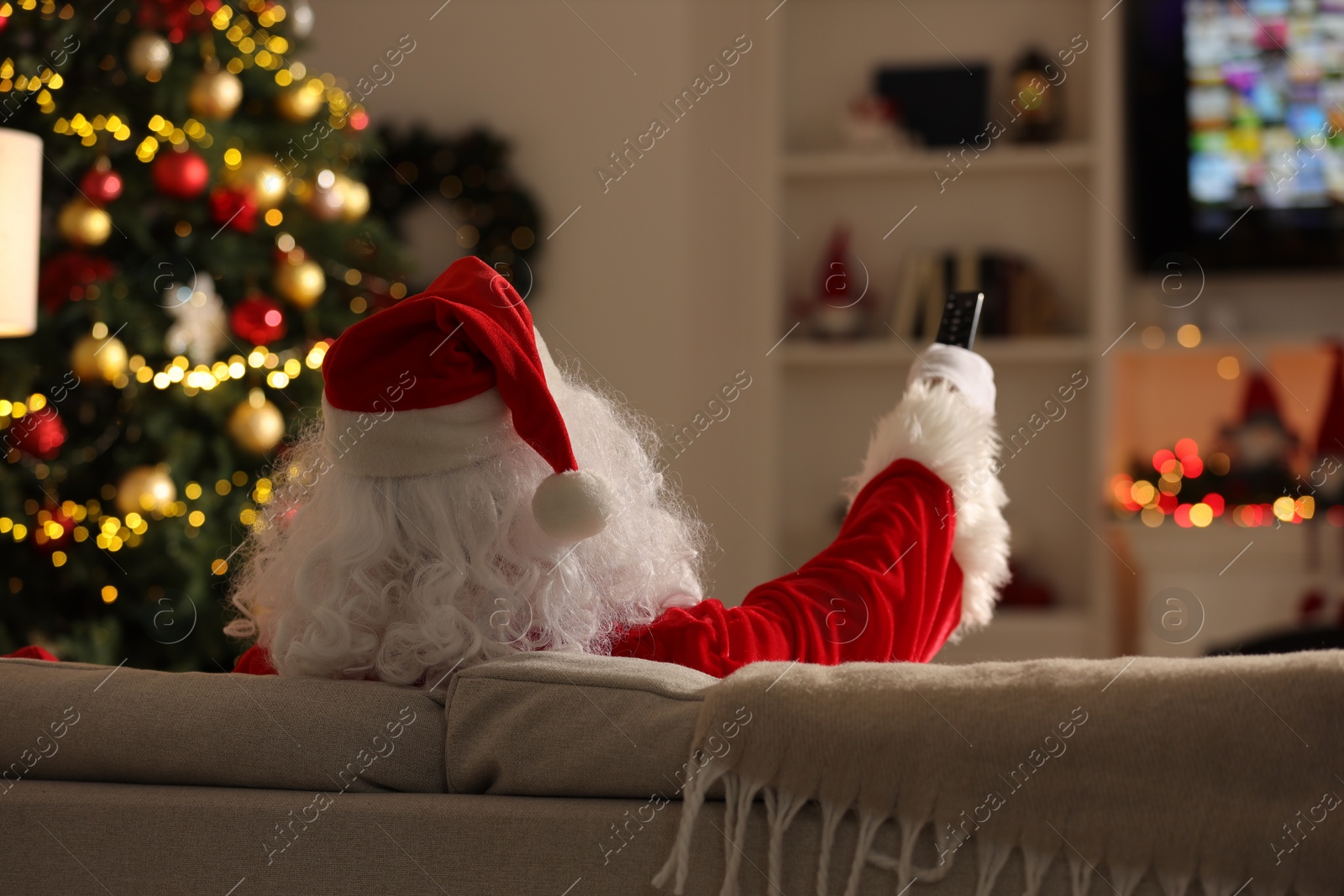 Photo of Merry Christmas. Santa Claus changing TV channels with remote control on sofa at home, back view