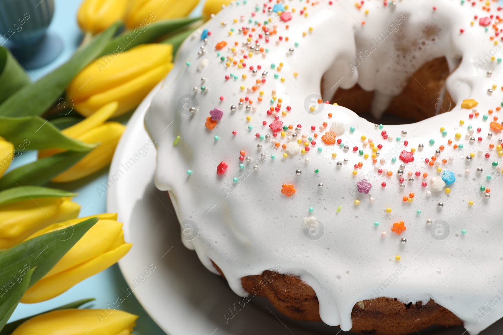 Photo of Delicious Easter cake decorated with sprinkles near beautiful tulips on light blue background, closeup