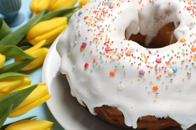 Delicious Easter cake decorated with sprinkles near beautiful tulips on light blue background, closeup