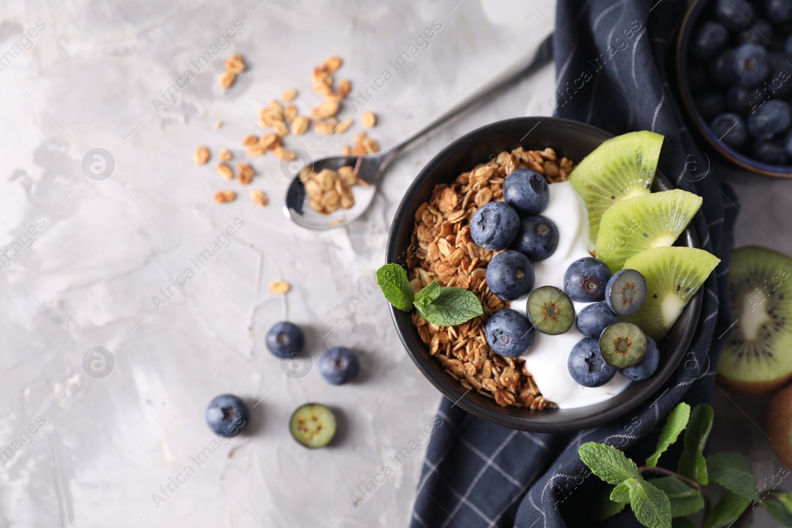 Photo of Tasty granola with yogurt, blueberries and kiwi in bowl on gray textured table, flat lay. Space for text