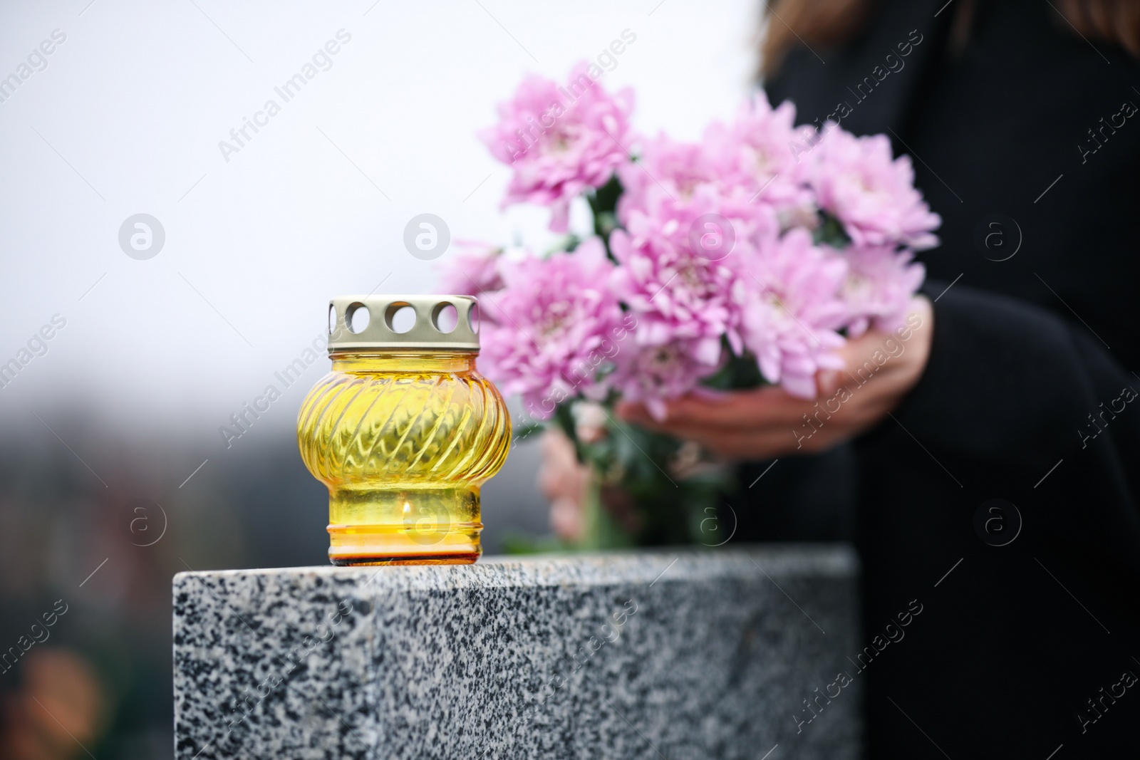 Photo of Woman holding chrysanthemum flowers near grey granite tombstone with candle outdoors, closeup. Funeral ceremony