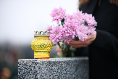 Photo of Woman holding chrysanthemum flowers near grey granite tombstone with candle outdoors, closeup. Funeral ceremony