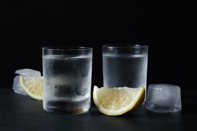 Photo of Shot glasses of vodka with lemon slices and ice on black background