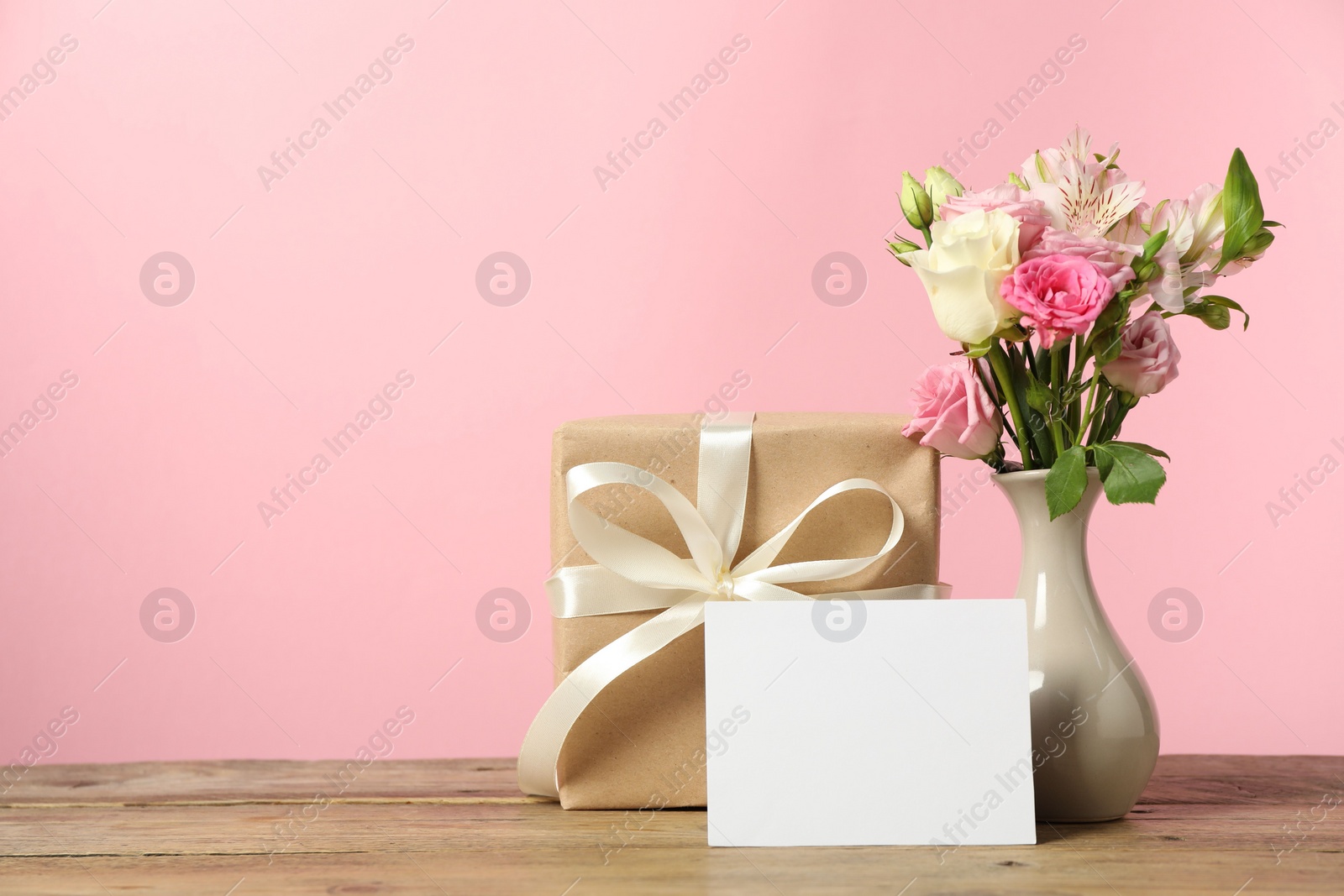 Photo of Happy Mother's Day. Gift box, blank card and bouquet of beautiful flowers in vase on wooden table against pink background. Space for text