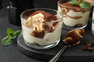 Photo of Delicious tiramisu in glasses, spoon, coffee beans and mint leaves on black table, closeup