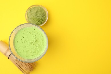 Glass of tasty matcha smoothie, powder and bamboo whisk on yellow background, flat lay. Space for text