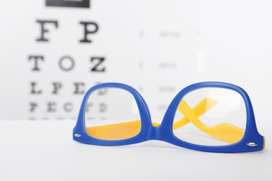Photo of Glasses with corrective lenses on table against eye chart
