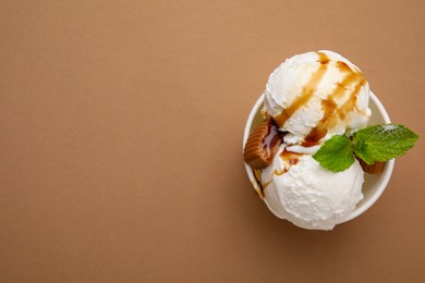 Scoops of tasty ice cream with caramel sauce, mint and candies in paper cup on light brown background, top view. Space for text