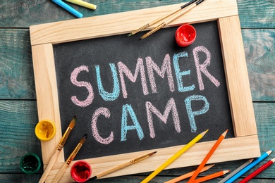 Photo of Text "SUMMER CAMP" on chalkboard, brushes and paints, top view