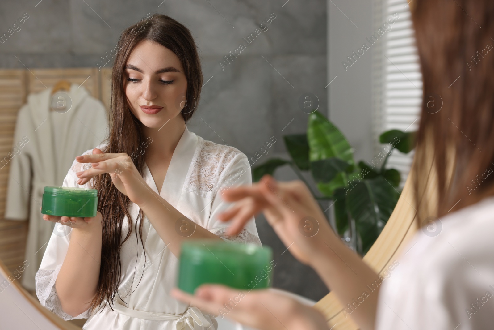 Photo of Young woman holding jar of aloe hair mask near mirror in bathroom