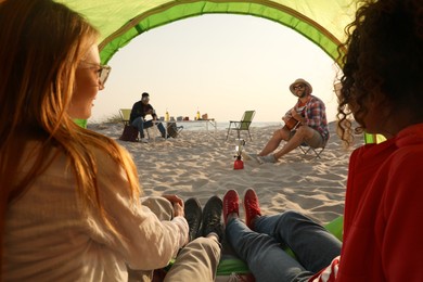 Photo of Friends resting on sandy beach. View from camping tent