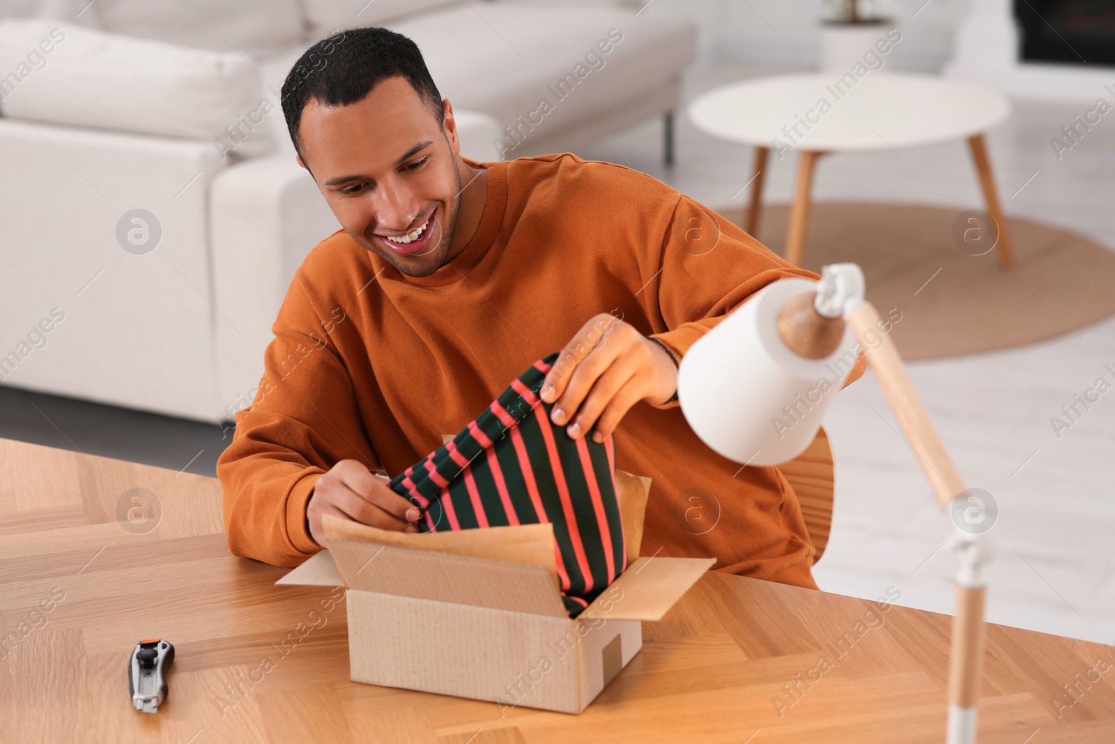 Photo of Happy young man opening parcel at table indoors. Internet shopping