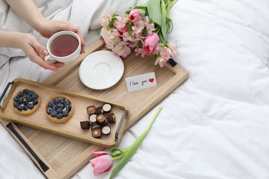 Photo of Tasty breakfast served in bed. Woman with tea, desserts, flowers and I Love You card at home, above view. Space for text