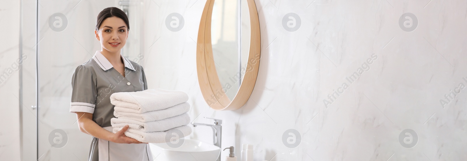 Image of Young chambermaid holding stack of fresh towels in bathroom, space for text. Banner design