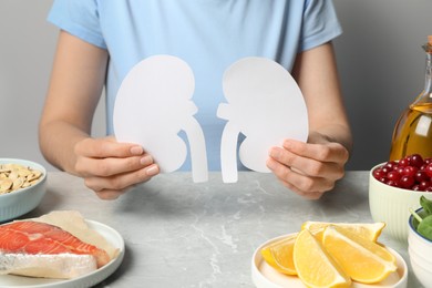 Photo of Woman holding paper cutout of kidneys near different products on light grey table, closeup