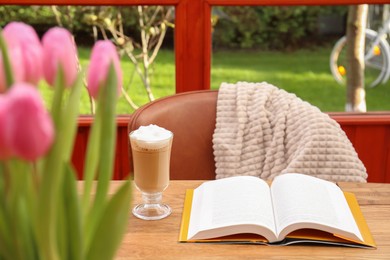Glass of delicious cocoa, book on wooden table and chair with blanket at terrace