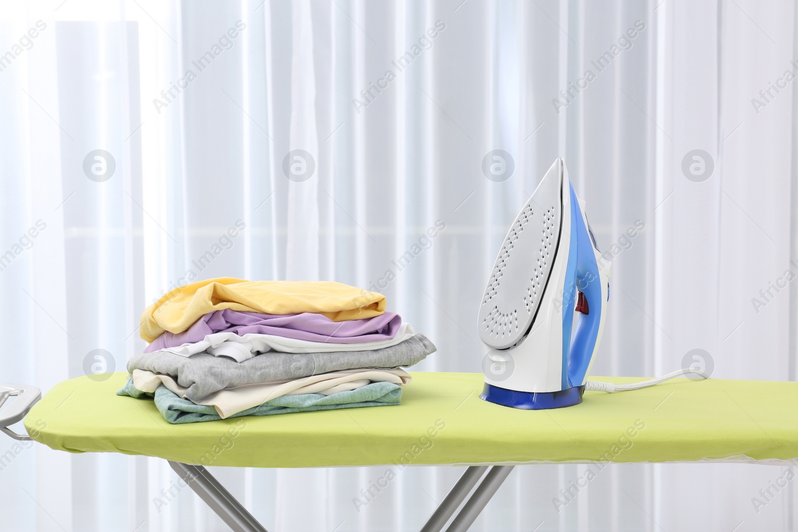 Photo of Board with modern iron and stack of clothes indoors. Laundry day